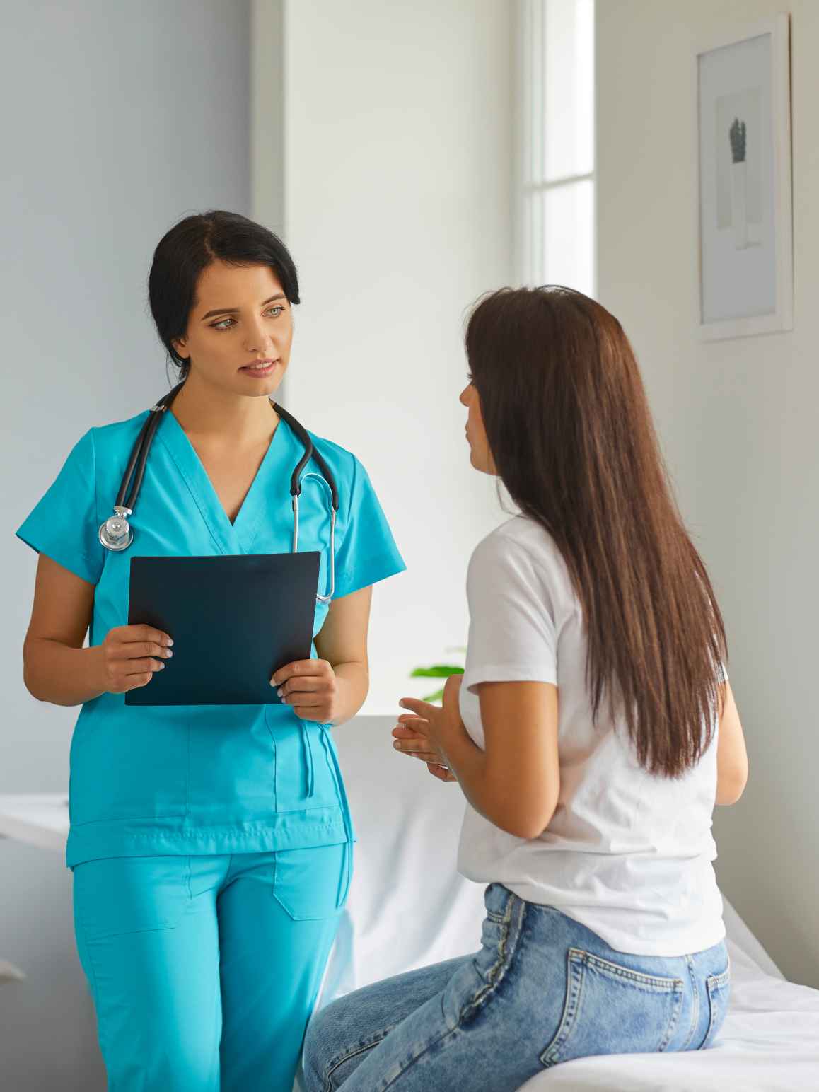 A healthcare professional in bright turquoise scrubs stands holding a clipboard while talking to a patient sitting on an examination table in a bright clinic room.