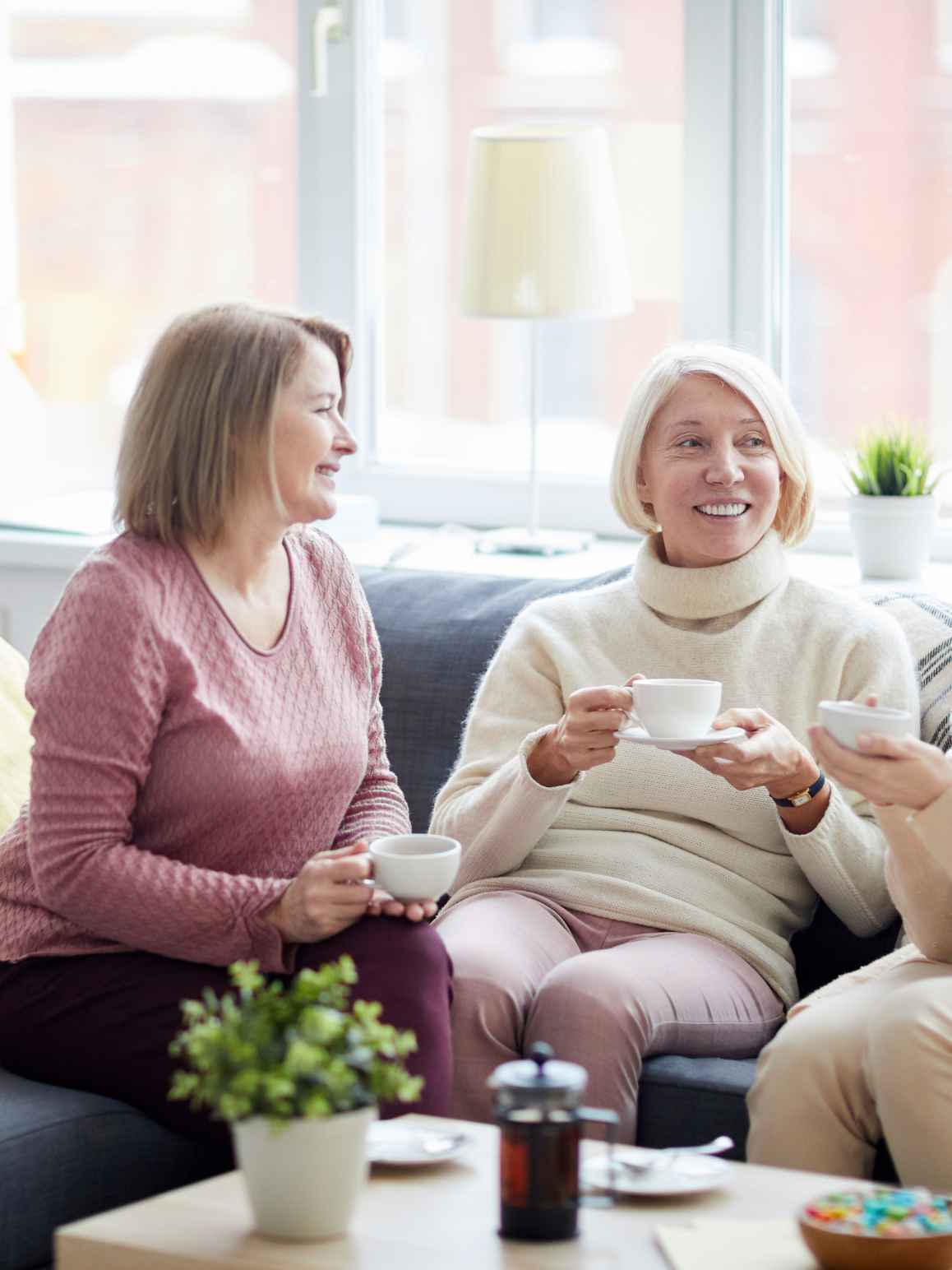 Two women sitting on a couch in a bright living room, smiling and holding white teacups. One wears a textured dusty rose sweater and dark pants, and the other wears a cream turtleneck sweater and pink pants.